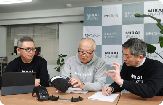 Three men sitting at table holding Mirai Speaker prototype